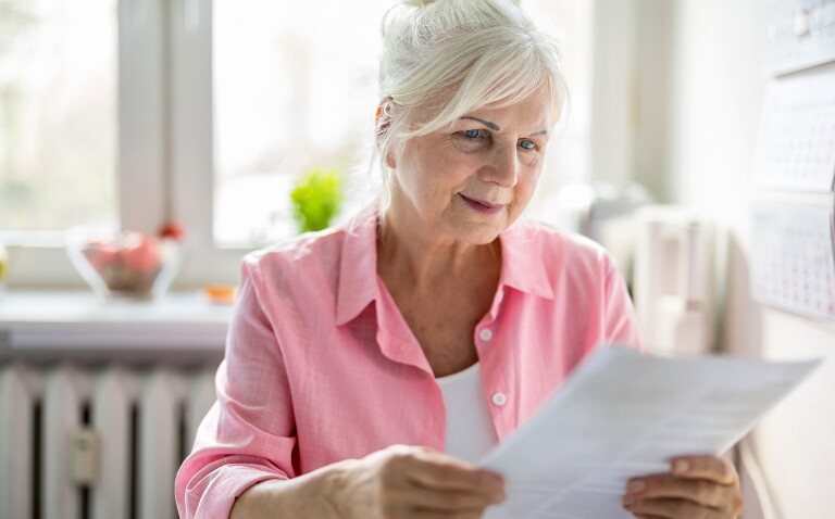A woman wearing a pink shirt and white tshirt sat at a kitchen table and reading a letter to illustrate a mammogram invitation