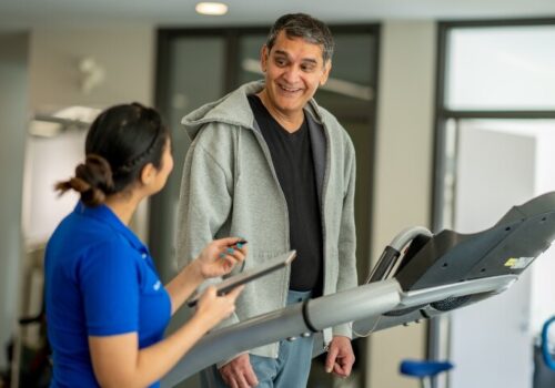 A senior gentleman walking on a treadmill in a gym during a pulmonary rehabilitation appointment and smiling at female healthcare worker who is standing beside him as she guides his exercise regimen and takes notes to illustrate a new framework from the BTS