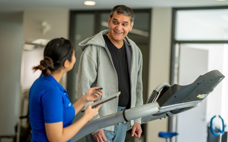 A senior gentleman walking on a treadmill in a gym during a pulmonary rehabilitation appointment and smiling at female healthcare worker who is standing beside him as she guides his exercise regimen and takes notes to illustrate a new framework from the BTS