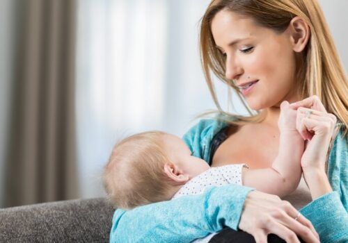 A woman wearing a blue cardigan sitting on a grey sofa and breastfeeding her baby to illustrate the finding that human milk-gut microbiome interactions are linked to childhood blood pressure