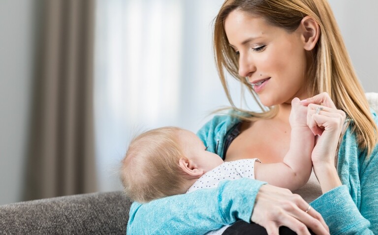 A woman wearing a blue cardigan sitting on a grey sofa and breastfeeding her baby to illustrate the finding that human milk-gut microbiome interactions are linked to childhood blood pressure