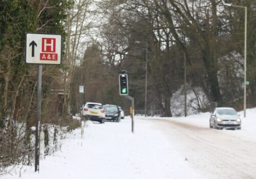A snowy street with cars left abandoned and a hospital road sign pointing forwards to A&E to illustrate the impact of temperature extremes on health and NHS resources and costs