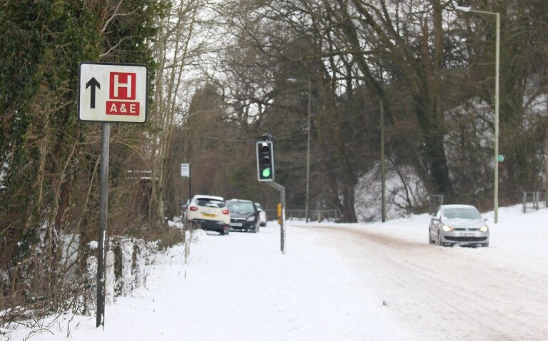 A snowy street with cars left abandoned and a hospital road sign pointing forwards to A&E to illustrate the impact of temperature extremes on health and NHS resources and costs