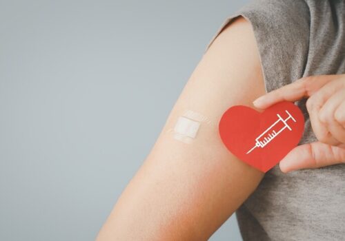 A close up of a woman holding a red heart with a drawing of a syringe on it in front of her arm with a plaster on it for a vaccination to illustrate the finding that maternal flu and Tdap vaccination is linked to fewer hospitalisations and emergency attendances in infants