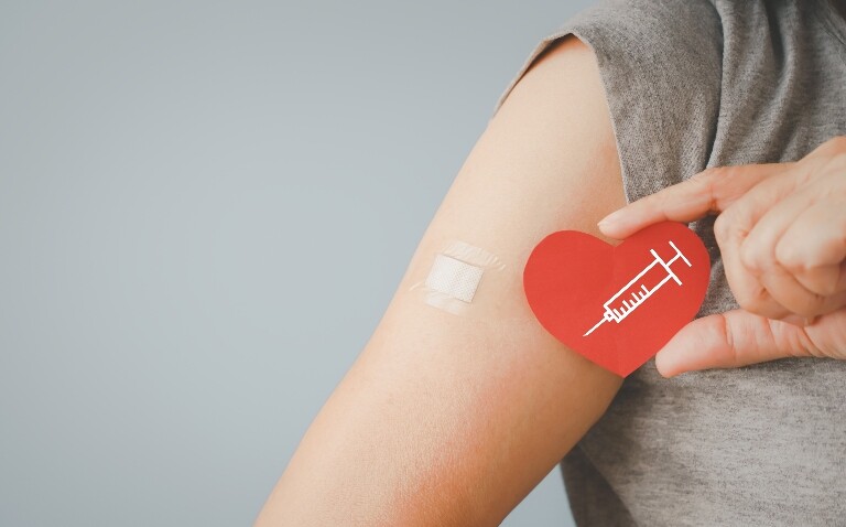 A close up of a woman holding a red heart with a drawing of a syringe on it in front of her arm with a plaster on it for a vaccination to illustrate the finding that maternal flu and Tdap vaccination is linked to fewer hospitalisations and emergency attendances in infants