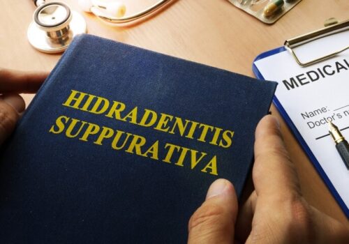 A book with title Hidradenitis Suppurativa on a table with a medical history form on a clipboard behind it and a stethoscope and a blister pack of medicines above it and a doctor's hands holding each side to illustrate quality-approved patient-reported outcome measures being identified for HS