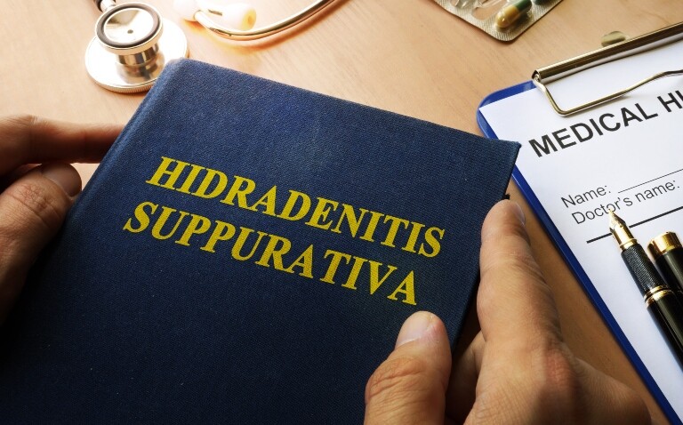 A book with title Hidradenitis Suppurativa on a table with a medical history form on a clipboard behind it and a stethoscope and a blister pack of medicines above it and a doctor's hands holding each side to illustrate quality-approved patient-reported outcome measures being identified for HS