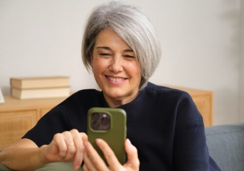 Middle aged woman sitting on a sofa and smiling while using a smartphone to illustrate robust evidence for the Axia app-based intervention in axial spondyloarthritis (axSpA)