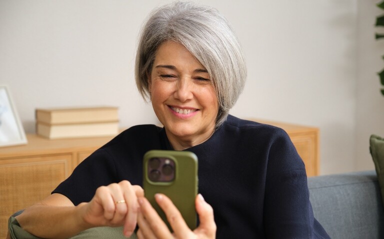 Middle aged woman sitting on a sofa and smiling while using a smartphone to illustrate robust evidence for the Axia app-based intervention in axial spondyloarthritis (axSpA)