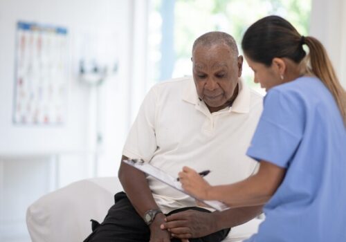 Elderly man sitting on an exam table while a healthcare professional in blue scrubs reviews paperwork with him