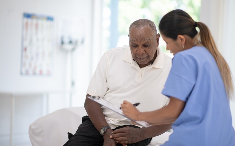 Elderly man sitting on an exam table while a healthcare professional in blue scrubs reviews paperwork with him