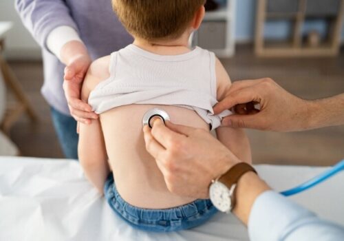 A young boy sat on a bed with his mum standing in front of him while a doctor holds a stethoscope to his back to illustrate valved holding chamber design and outcomes in young children with acute wheezing