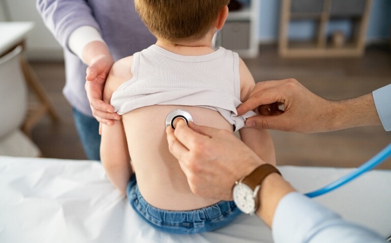 A young boy sat on a bed with his mum standing in front of him while a doctor holds a stethoscope to his back to illustrate valved holding chamber design and outcomes in young children with acute wheezing