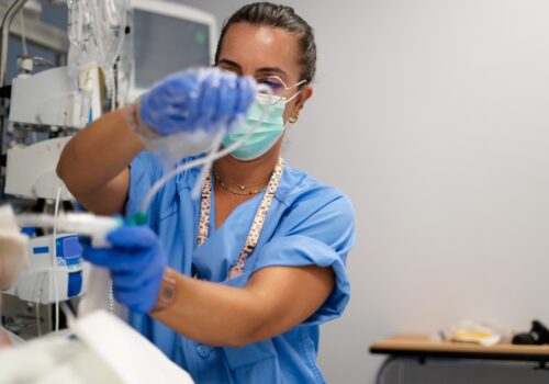 A medical professional wearing a surgical mask and working on a patient's ventilation system to illustrate calls for standardised infection prevention and risk assessment policies due to wide variation