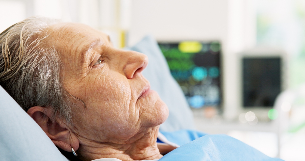 Close-up side profile of an elderly woman lying in a hospital bed with blurred medical monitors in the background to illustrate the exploration of invasive vs conservative strategies for frail older adults with NSTEMI