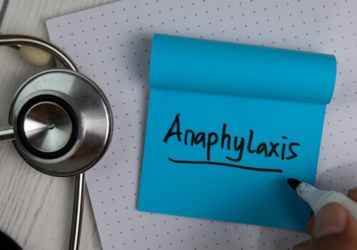 The word anaphylaxis being written on a blue post-it note on a desk next to a stethoscope