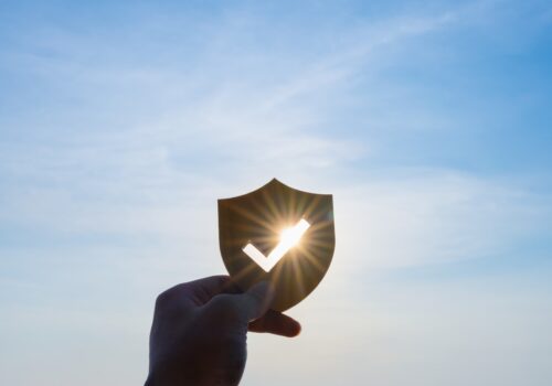 A hand holding a shield with a tick cut out of it against a blue sky with the sun coming through the middle to illustrate the prevention of SCC in immunocompromised patients via the SPOT-IT trial