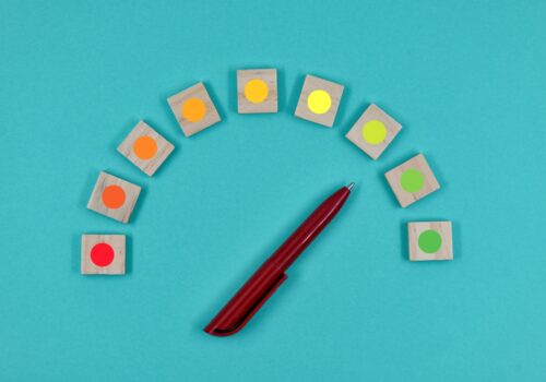 Wooden blocks with coloured dots arranged in a curved traffic-light scale from red to green with a burgundy pen pointing to the yellow to green transition on a turquoise background to illustrate the value of patient-reported outcomes during tildrakizumab psoriasis treatment